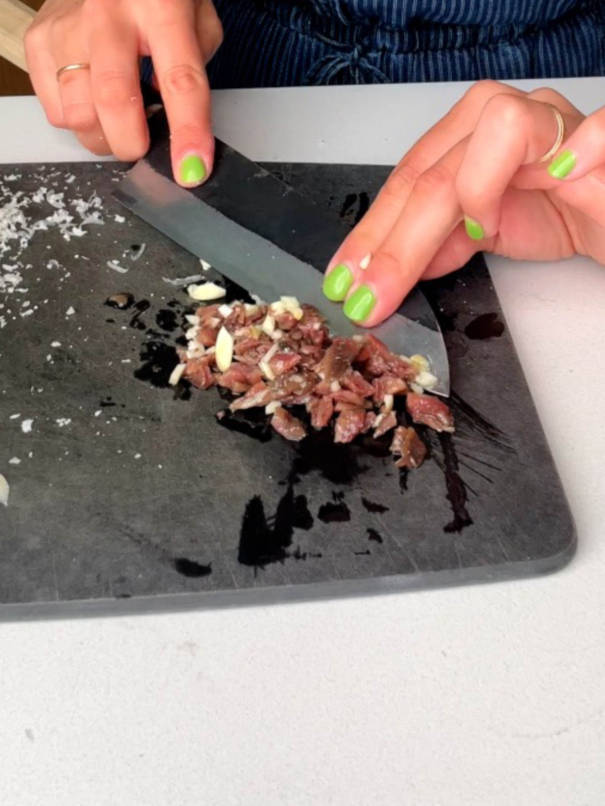 Mincing anchovies and garlic on a black cutting board with a knife and hands with green nail polish.