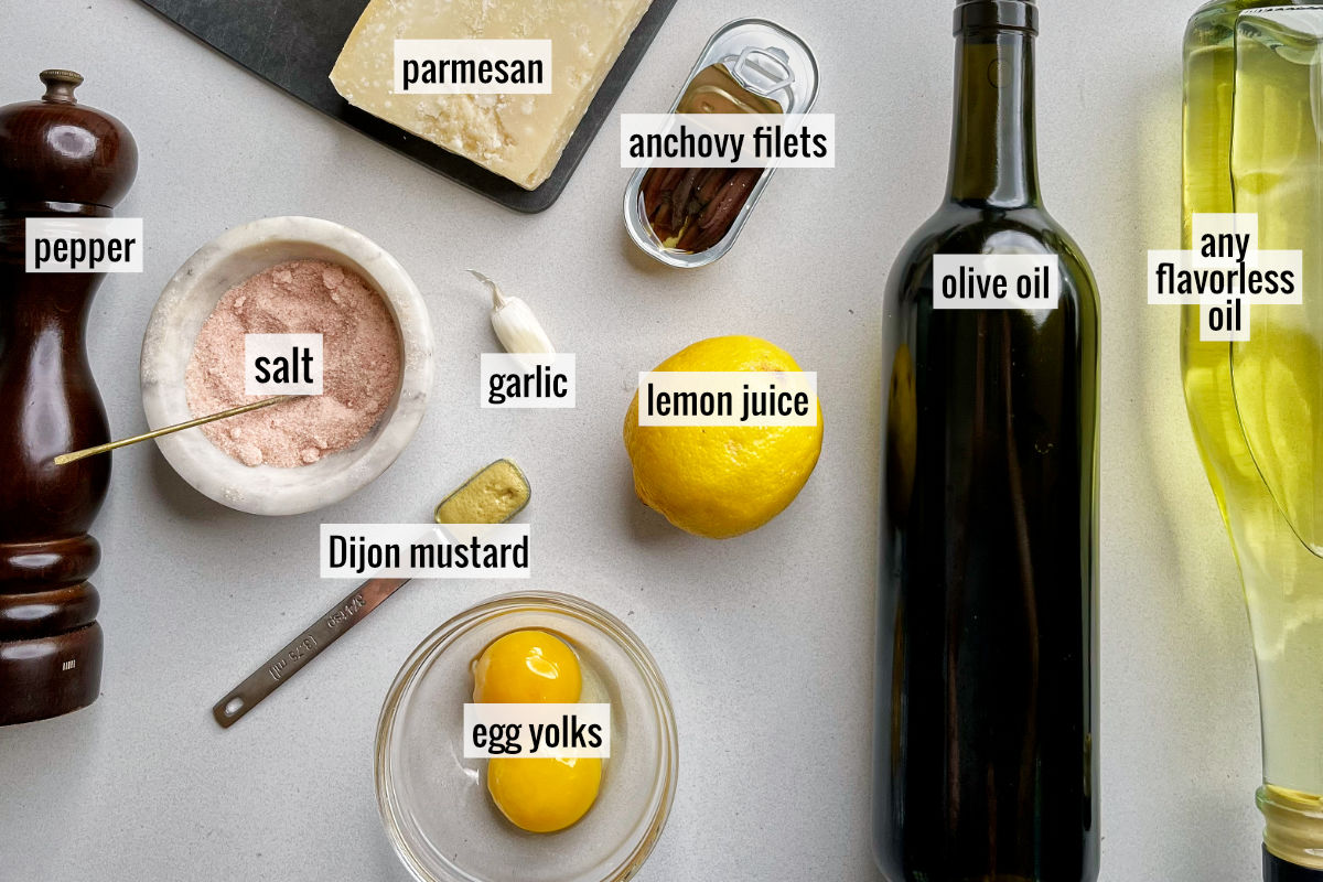 Ingredients laid out and labeled to make caesar salad dressing like anchovies, parmesan, oil, and lemon.