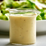 Caesar salad dressing in a glass beaker in front of a plate of lettuce.