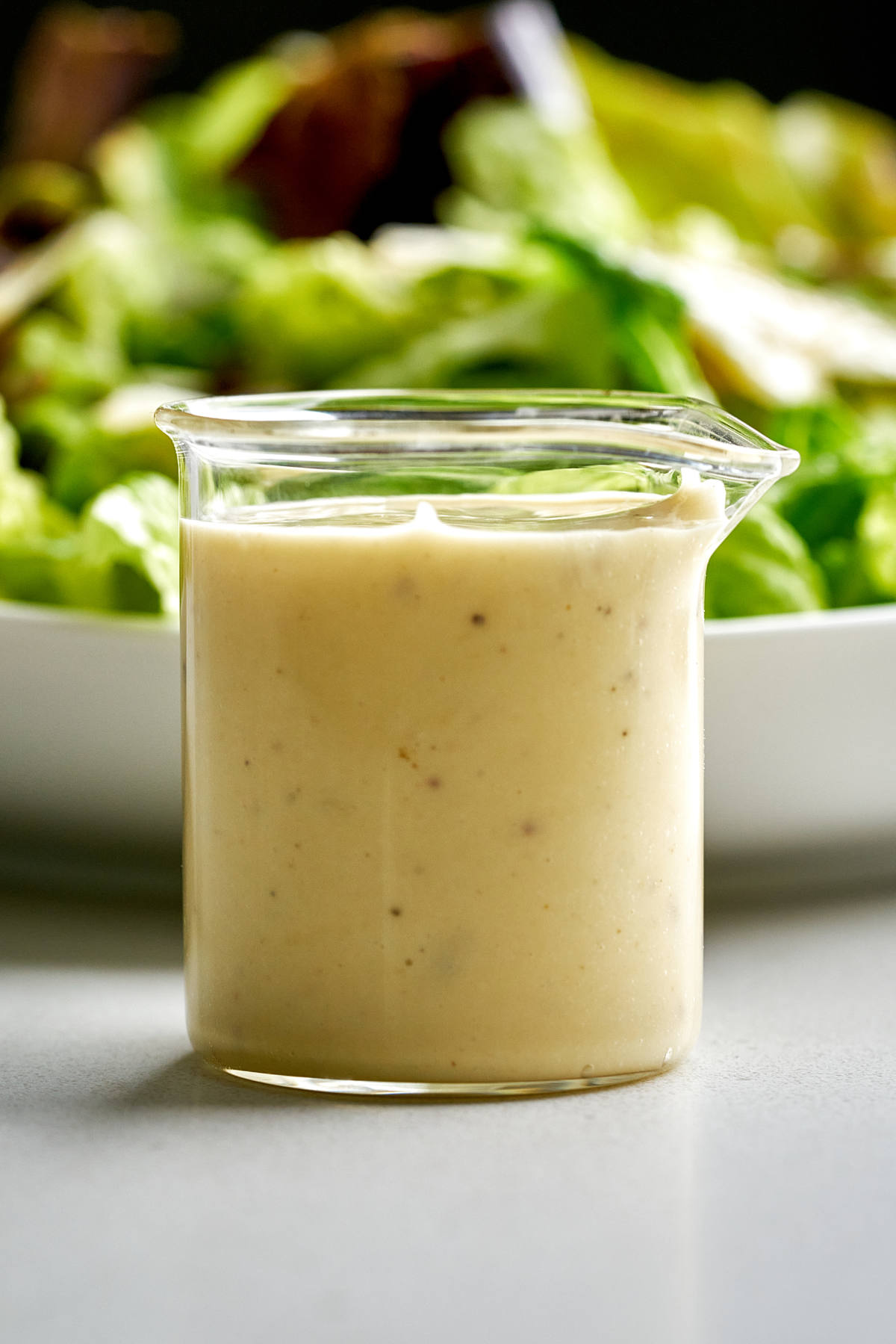 Caesar salad dressing in a glass beaker in front of a plate of lettuce.