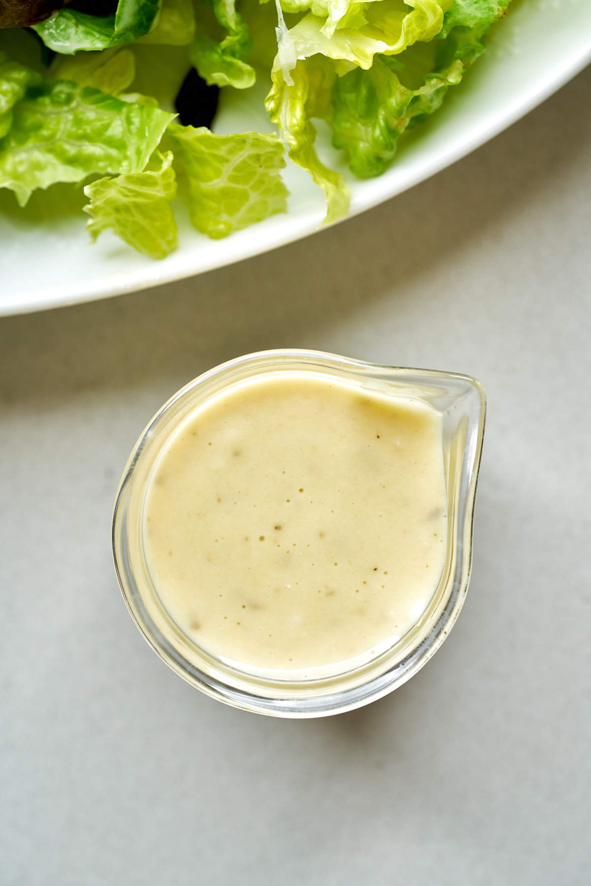 Top view of caesar salad dressing in a glass beaker next to a plate of lettuce.