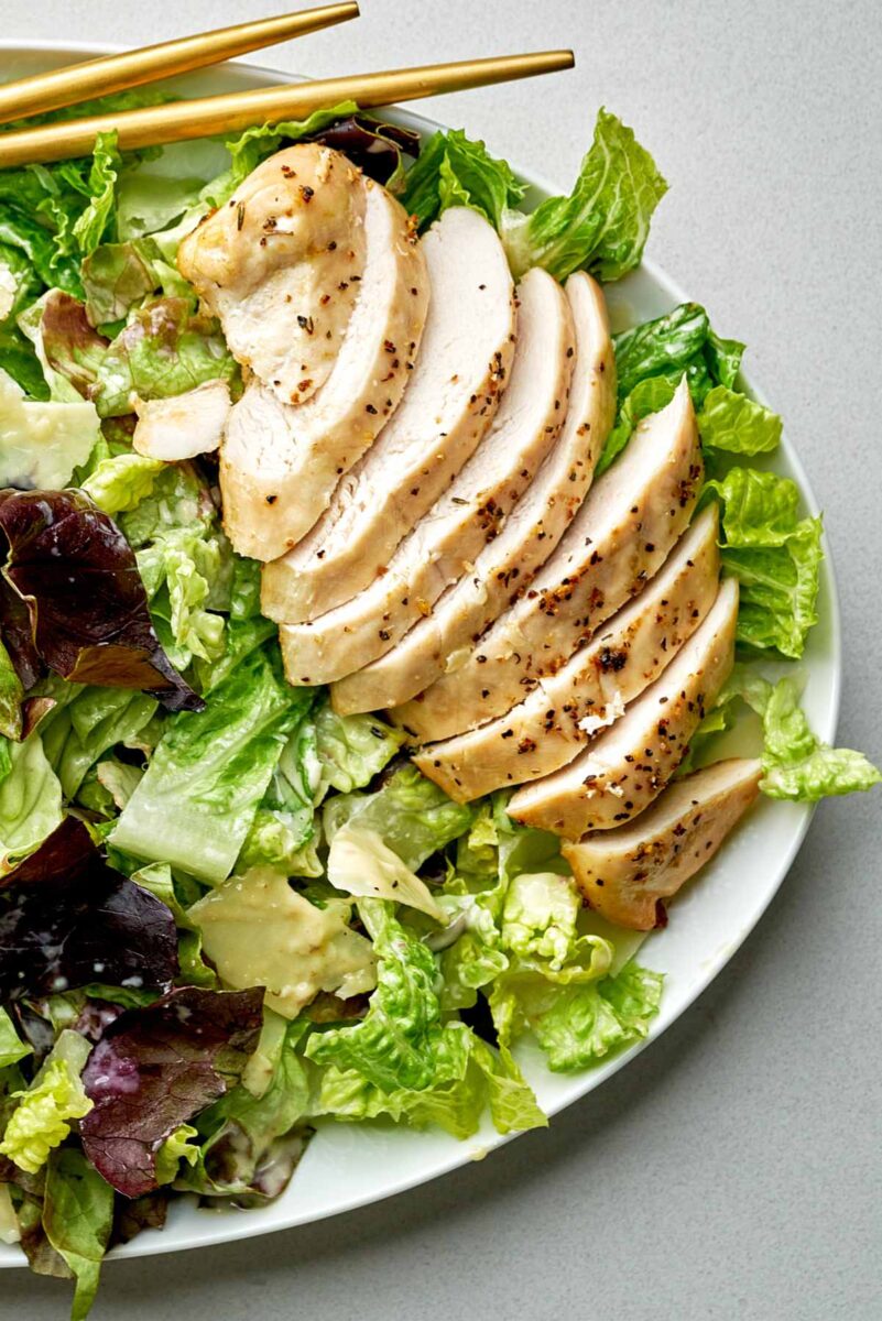 Sliced chicken breast on a bed of lettuce next to gold serving utensils.