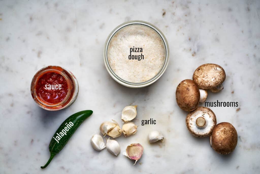 Top view of dough, pizza sauce, jalapeno, garlic, and mushrooms on a white countertop.