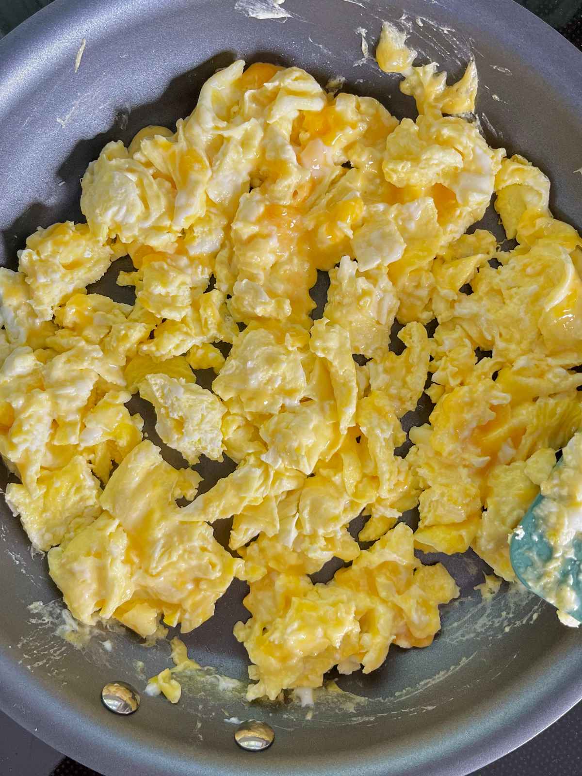 Scrambled eggs with bits of melted cheese in a dark grey non stick pan.