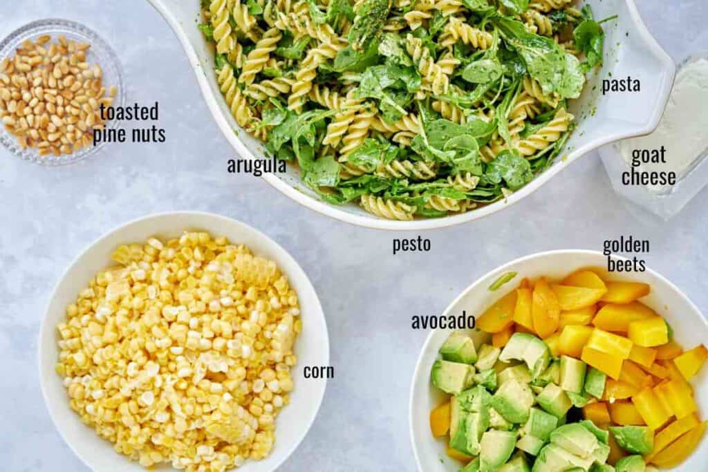 Ingredients for pasta salad including avocado, beets, corn, arugula, pesto, and noodles.