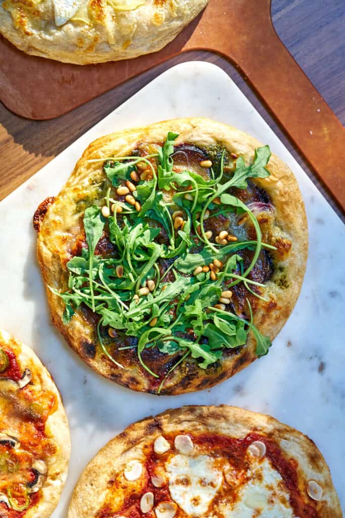 Pizza topped with arugula on white marble next to more pizza.