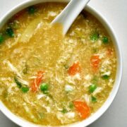 Bowl of yellow egg drop soup with carrots and peas.