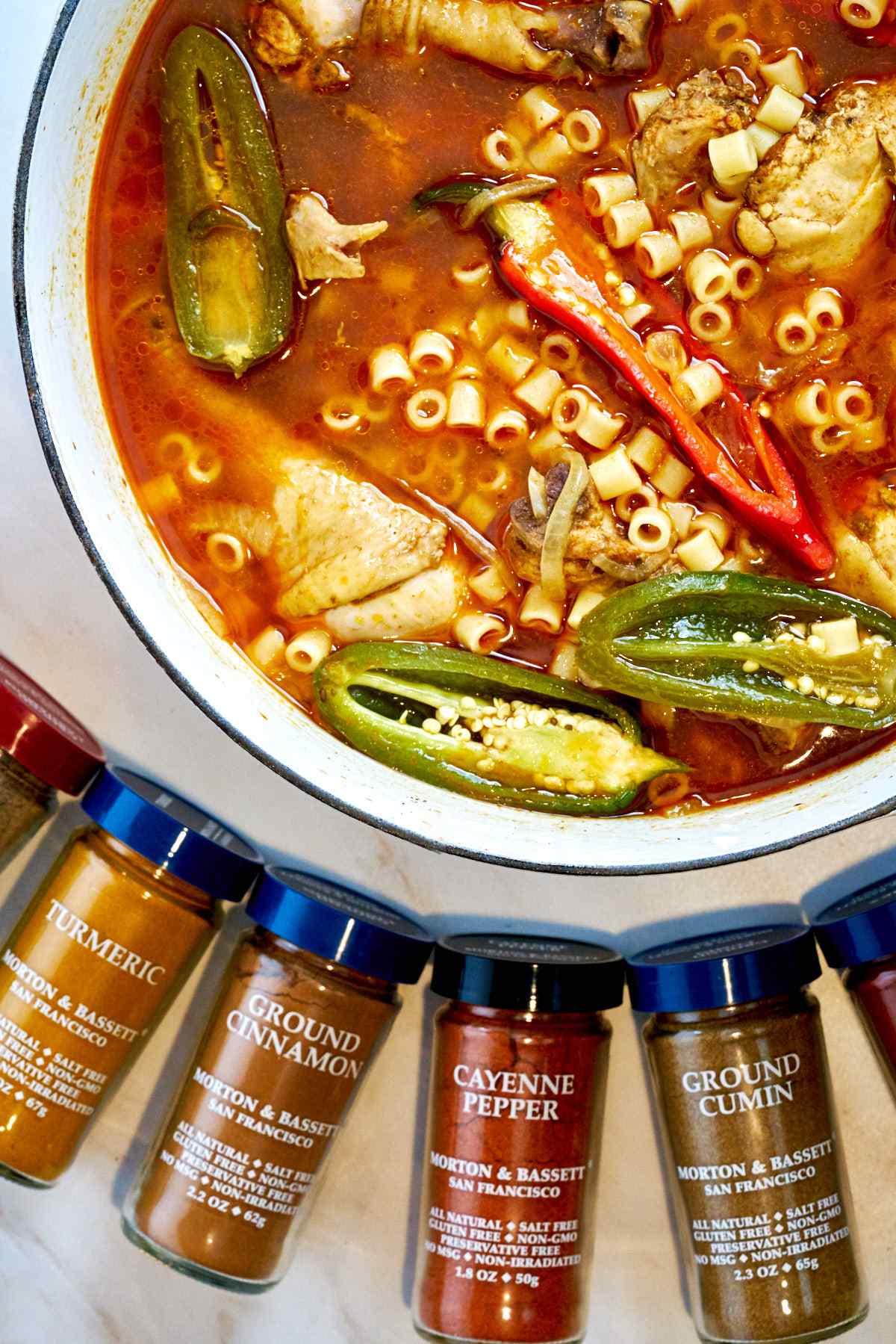 Pot of noodle soup with peppers next to spice bottles.