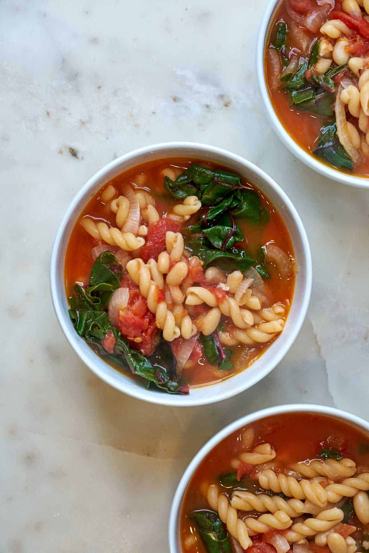 Three bowls of Bowl of red soup with noodles and greens.