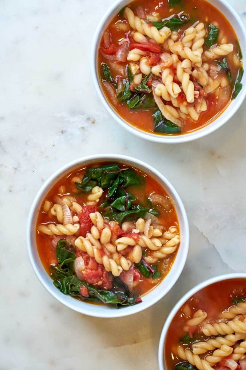Three bowls of Bowl of red soup with noodles and greens.