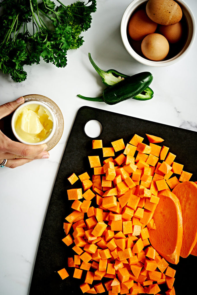 Sweet potato diced with eggs in a ramekin, butter, and green peppers on a marble backdrop.