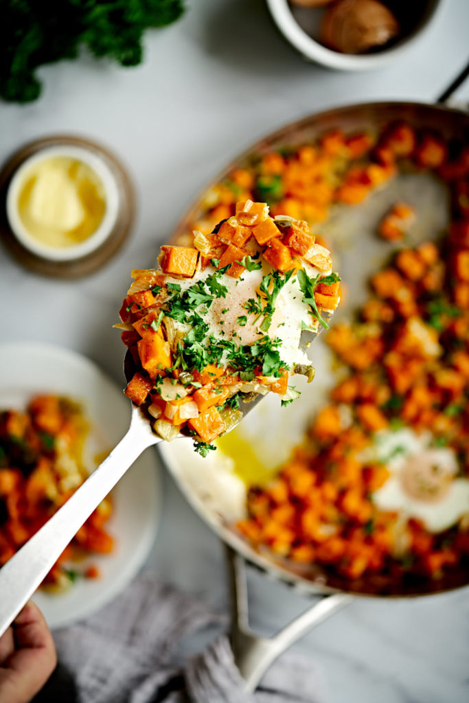 A close up of sweet potato hash on a silver spatula with egg and parsley.