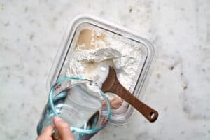 Pouring water into a container with flour, yeast, salt, and a wooden spoon.