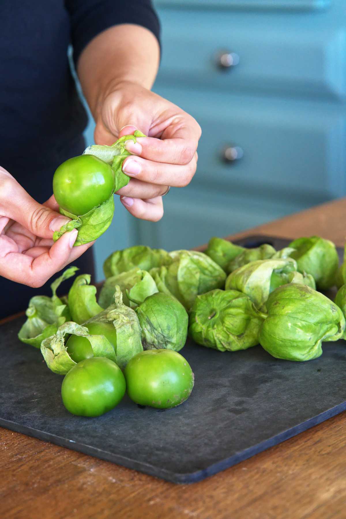 Woman peeling tomatillos on a black cutting board.