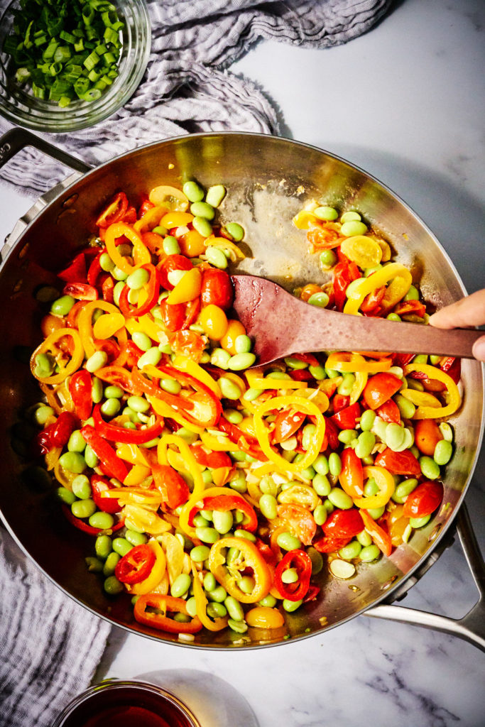 Fry pan with wooden spoon, tomatoes, peppers, and edamame.
