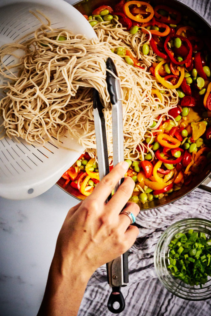 Pouring noodles into a stir fry from a colander with tongs.