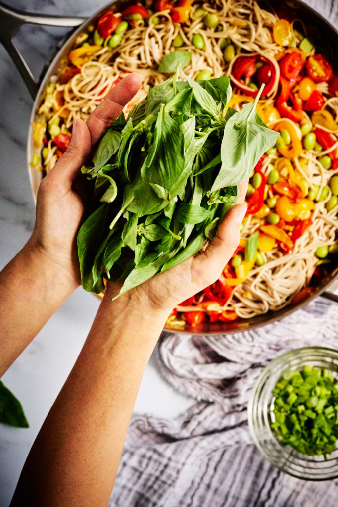 Two hands holding fresh basil over a fry pan with a noodle stir fry.