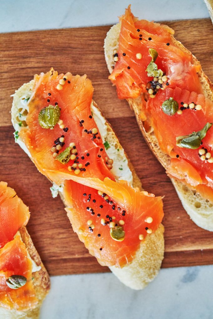 An overhead view of slices of bread arranged diagonally on a cutting board, topped with cream cheese and cured salmon.