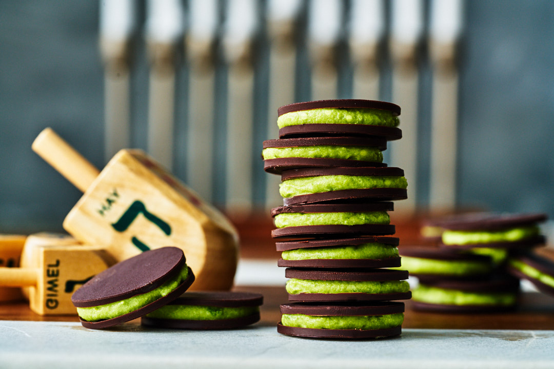 A stack of gelt layered with green filling next to dreidels and a chanukiyah.