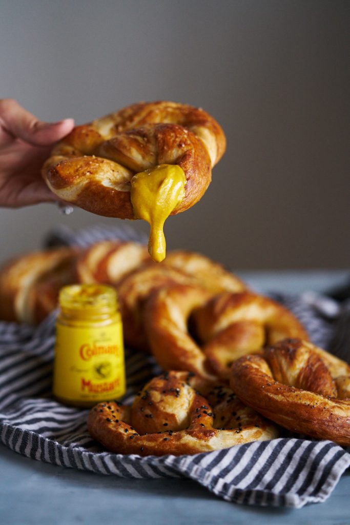 A hand holding a baked pretzel with mustard dripping off the edge.