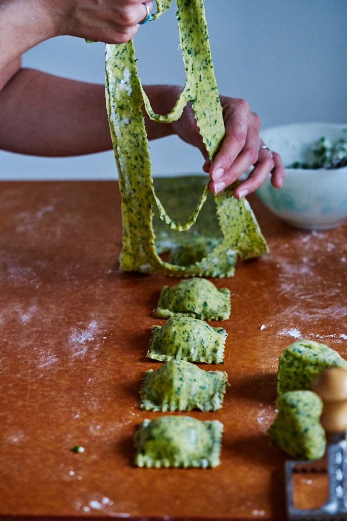 Hands lifiting speckled pasta up from square cut outs of ravioli on a floured counter.