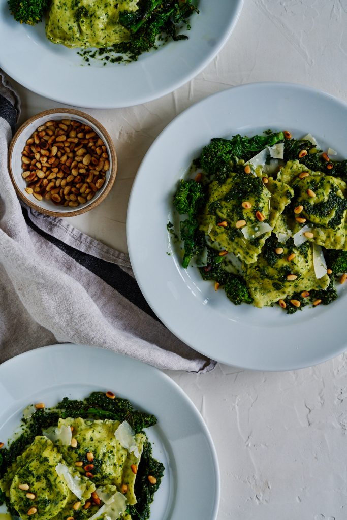 Three white plates with pesto-covered ravioli next to a small bowl od pine nuts.