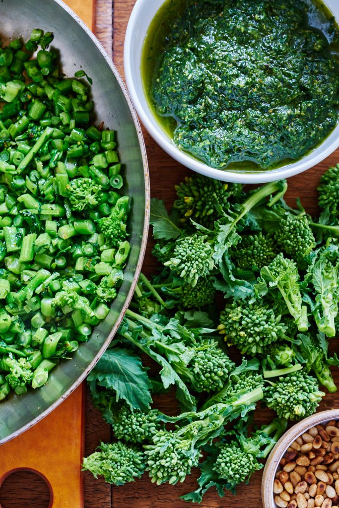 Chopped up broccoli rabe in a silver pan next to a bowl of green pesto and raw broccoli rabe.