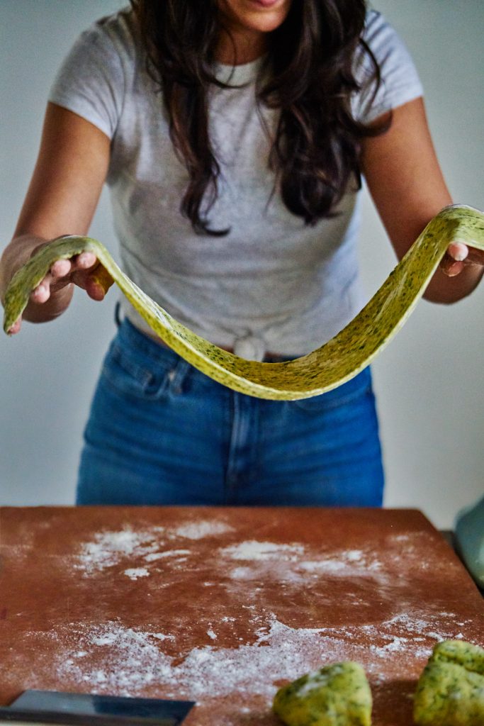 A woman stretching out a long peice of beige and green pasta above a floured counter.