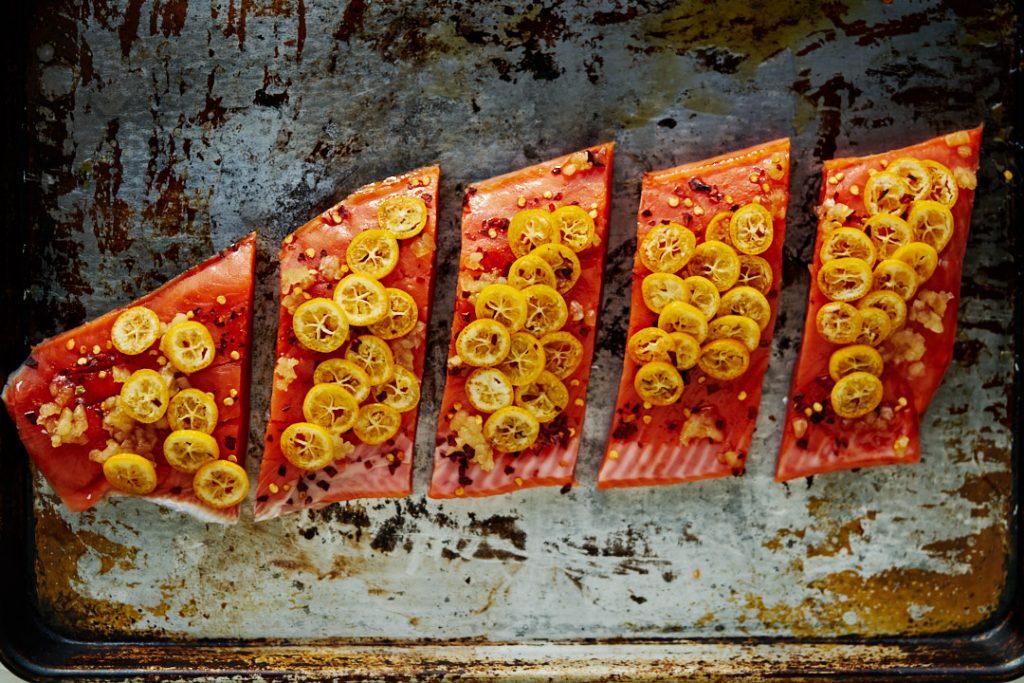 A large piece of salmon cut into steaks and covered in kumquat slices on a metal pan.