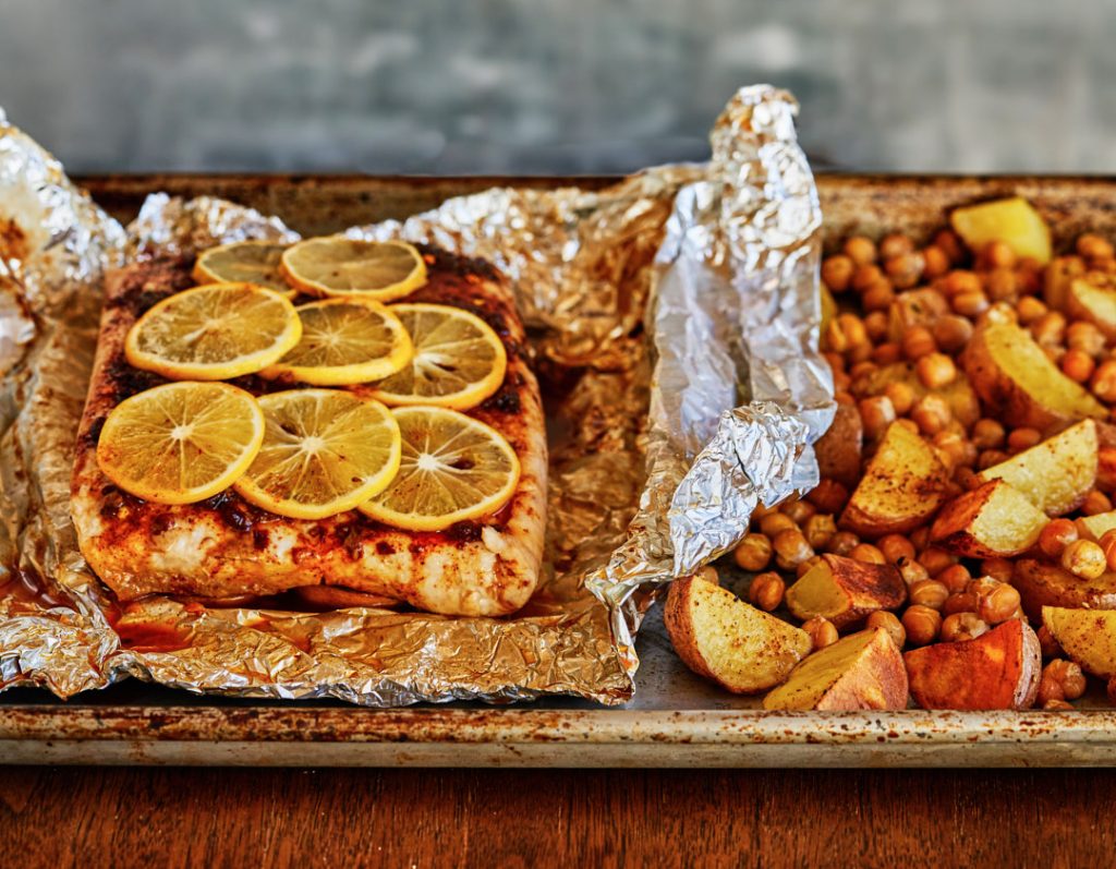 White fish with spices and lemon slices in foil, next to baked chick peas and potatoes in a metal pan.