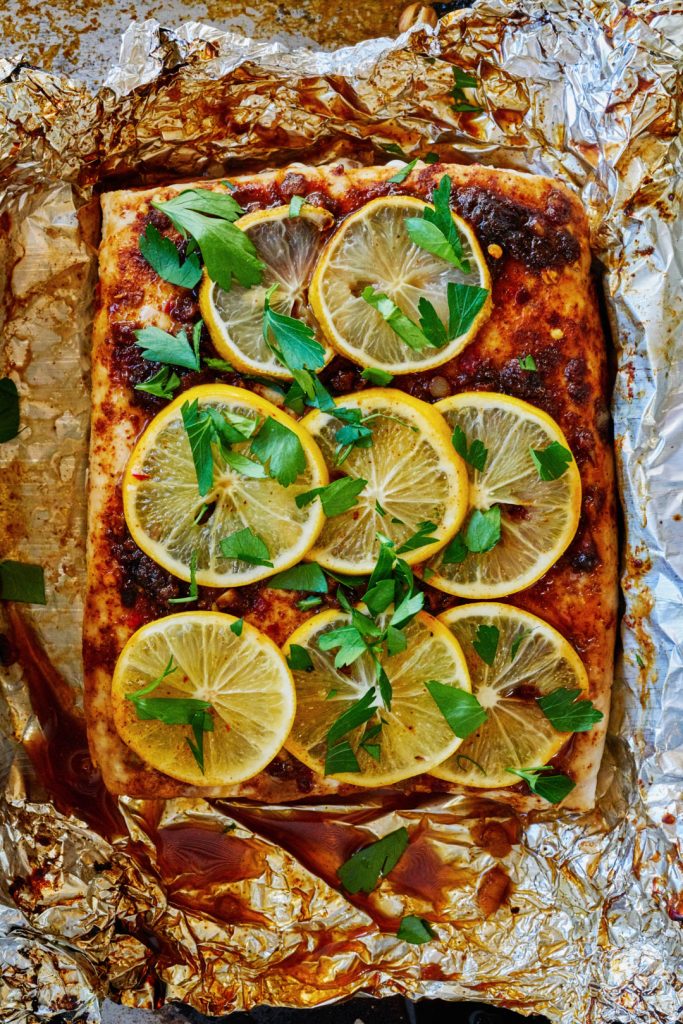 Close up of white fish on foil paper covered in spices, slices of lemon, and parsley.