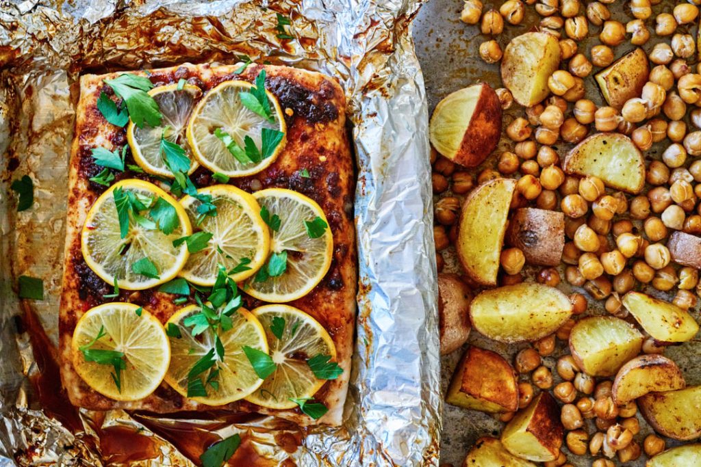 White fish with spices and lemon slices in a foil-lined pan, next to baked chick peas and potatoes.