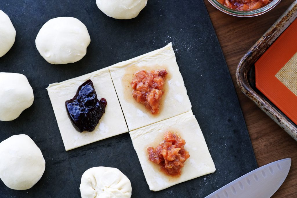 Squares of pastry with jan on top on a  black cutting board, with more balls of pastry beside it.