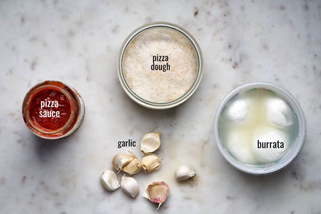 Top view of pizza sauce, dough, burrata, and garlic.