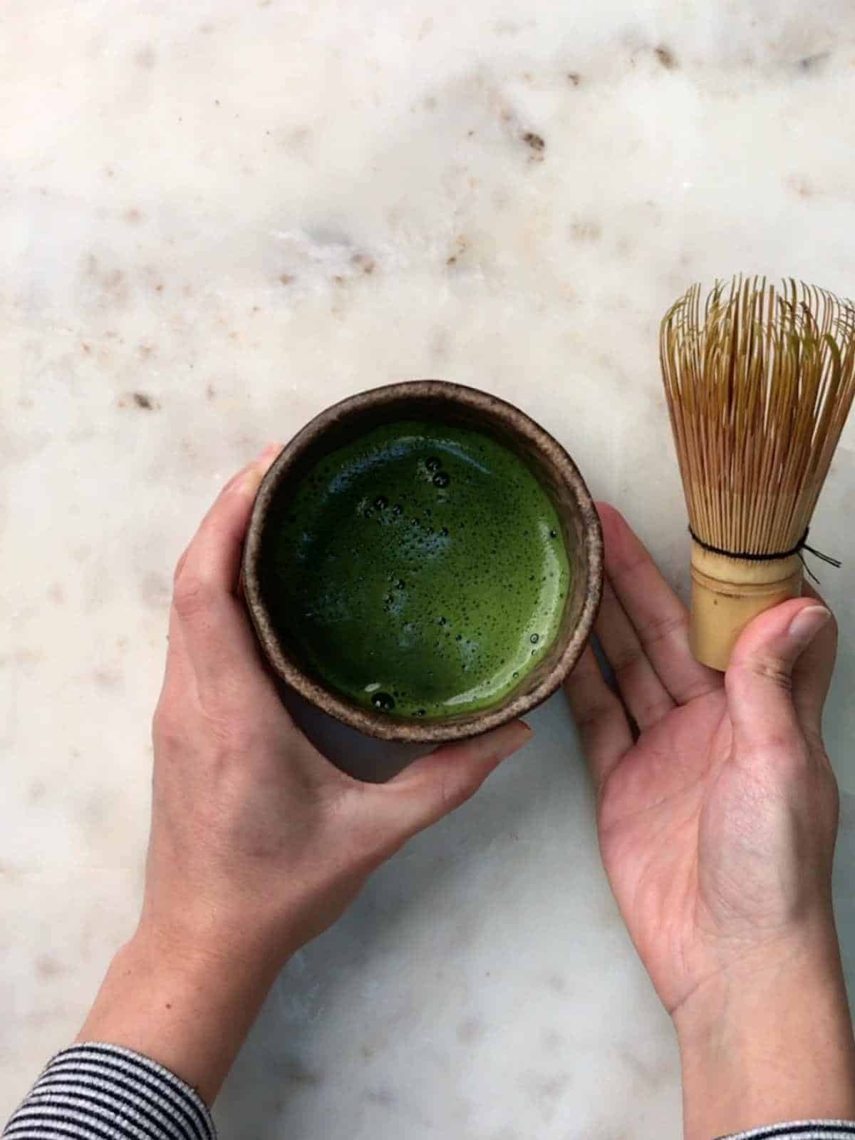 Two hands holding a ceramic cup of whisked green tea in a bowl.