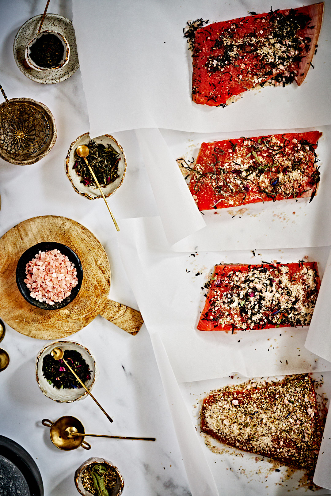 Small bowls of sugar, salt and tea leaves on a counter next to four salmon fillets coated in a salt, sugar, and tea mixture.