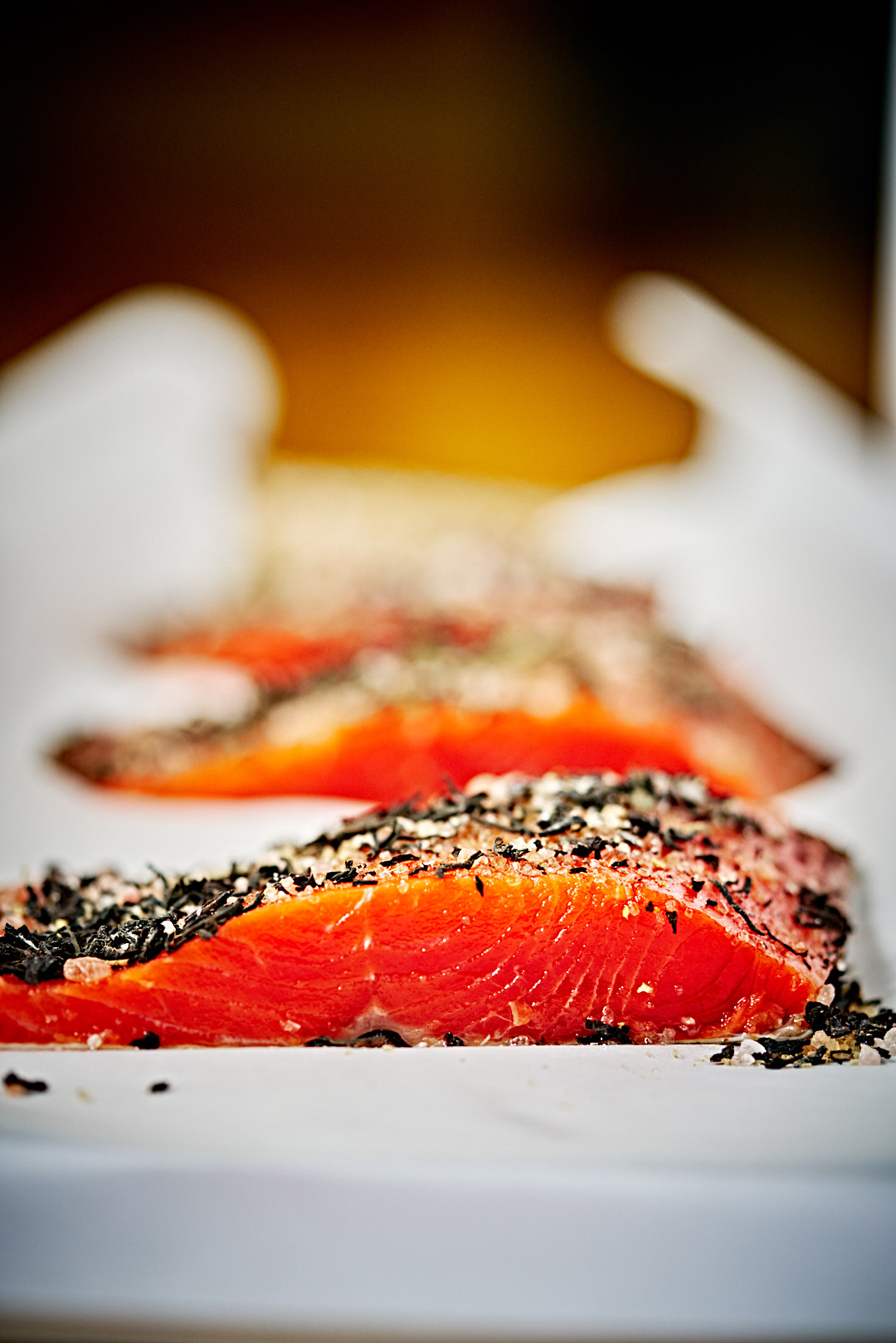 Sideways view of a orange salmon fillet covered in salt sugar and tea leaves on parchment paper.