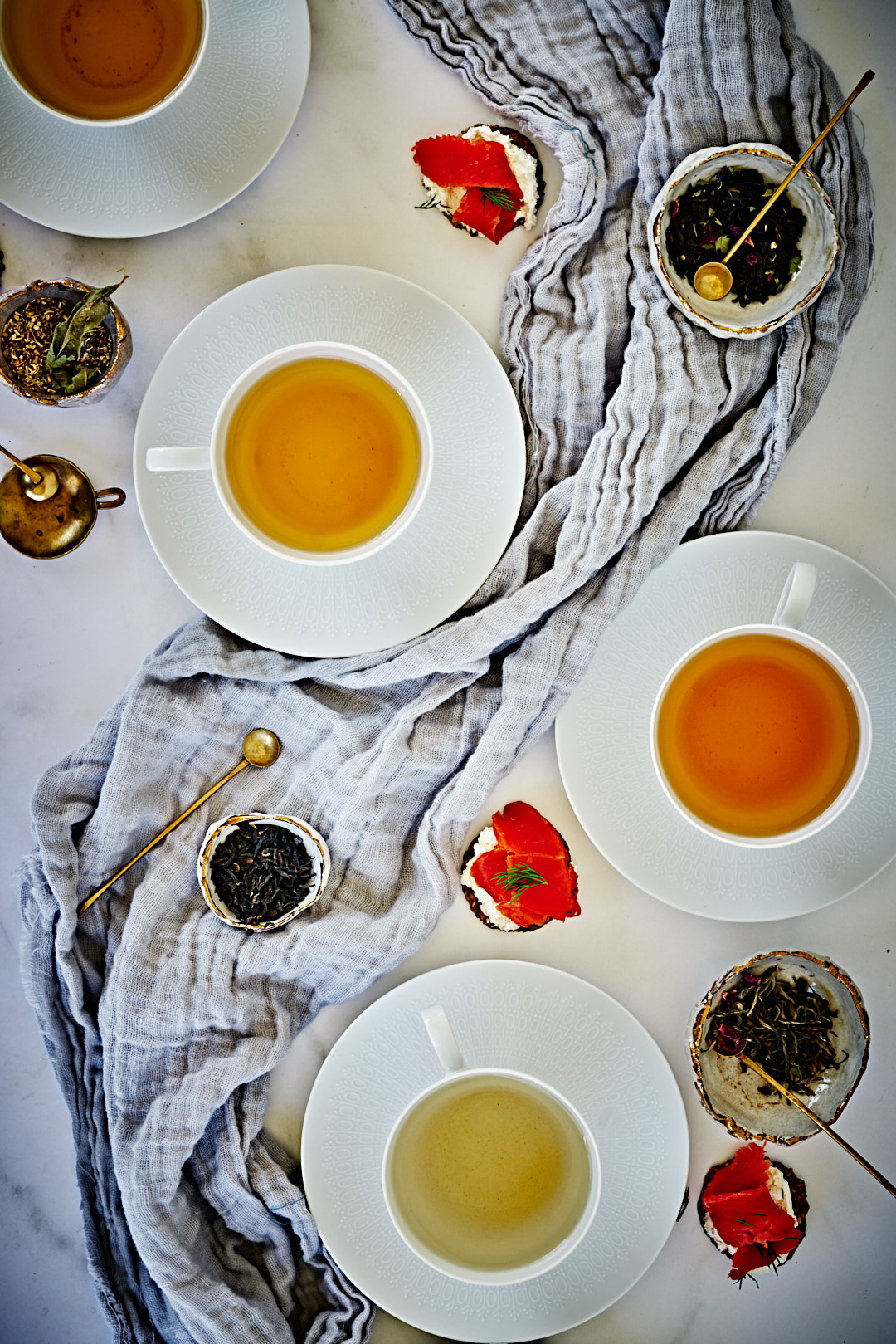 A flat lay of cups of tea, bowls of loose leaf tea, and sliced salmon on Cream cheese and bread.