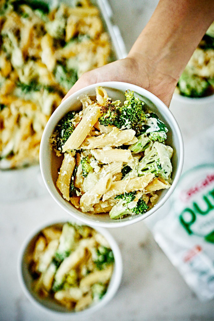 A hand holding a white bowl of penne pasta with broccoli and white sauce.