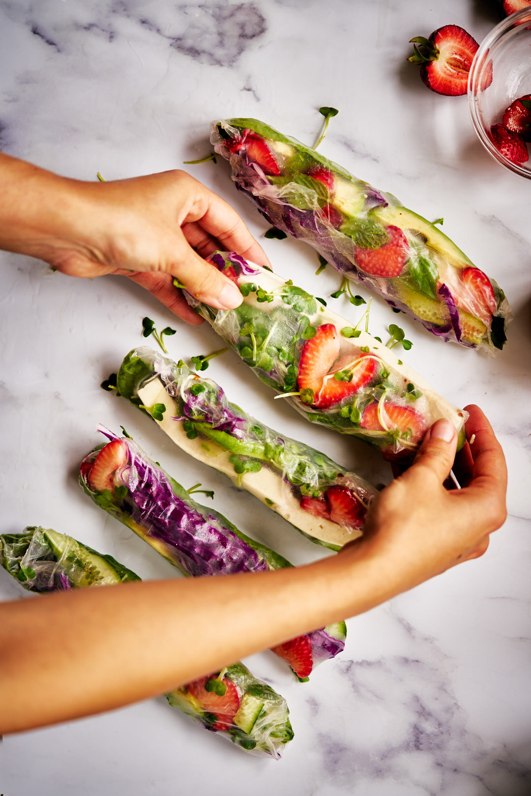 Flatlay of hands holding colorful tofu summer salad rolls on white marble surface.