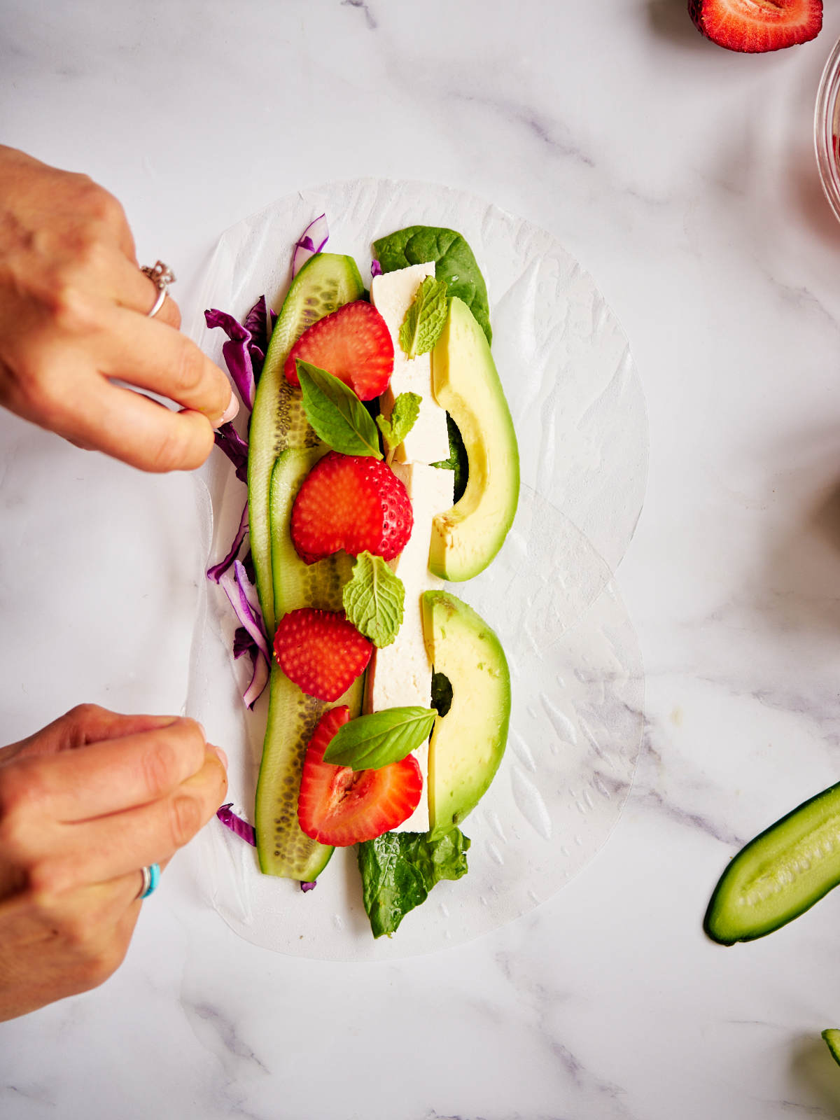 Two hands rolling a salad roll filled with avocado and strawberries.