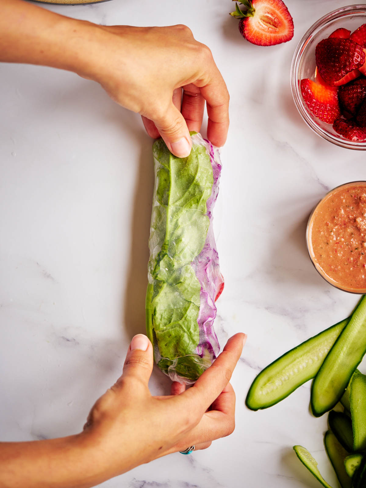 Two hands rolling a salad roll next to cucumbers, dressing, and strawberries.