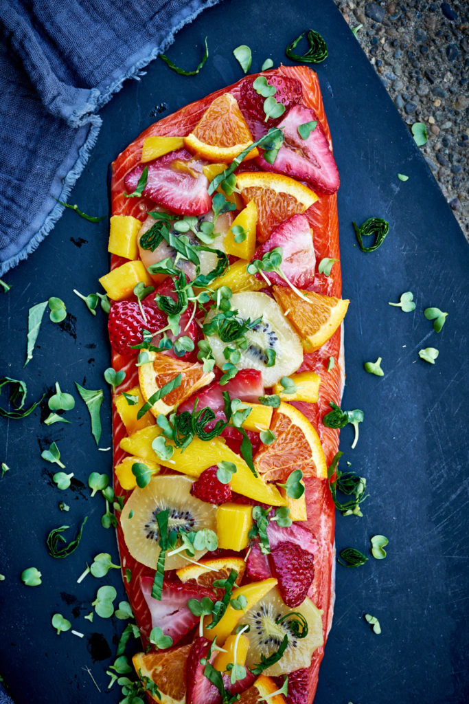 Salmon topped with summer fruit and fresh herbs on a cutting board.