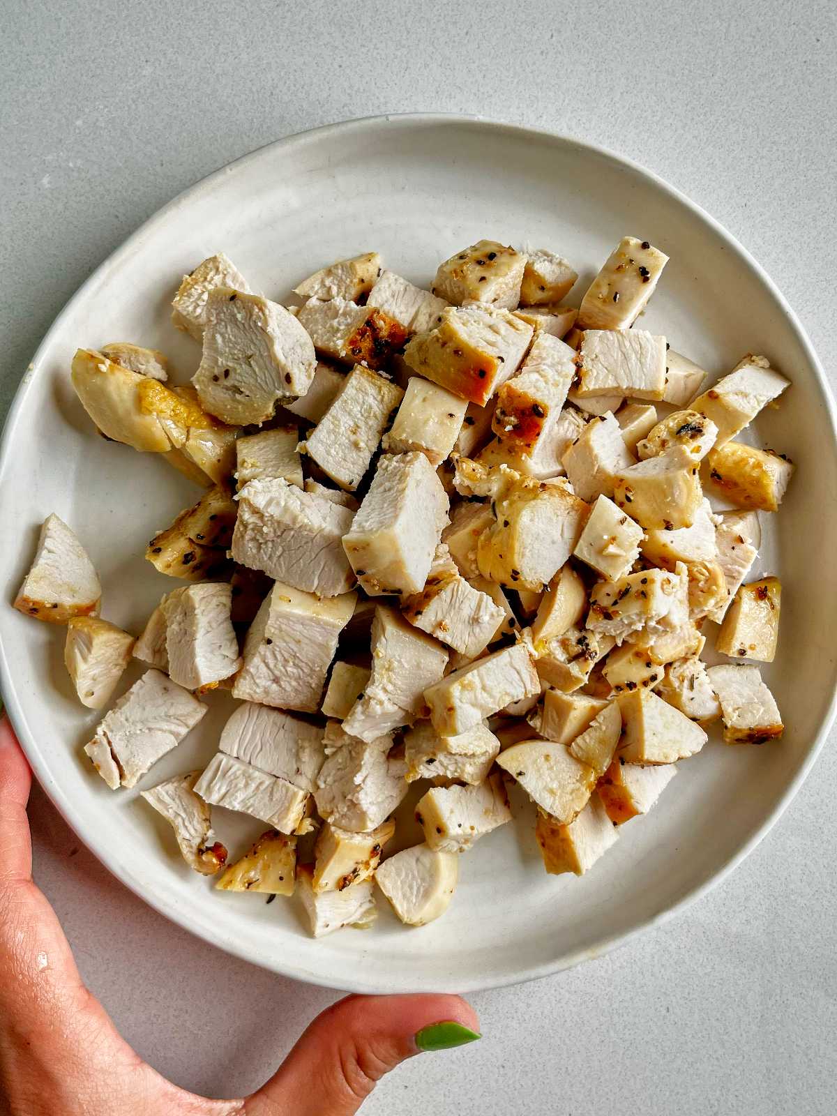 Cubed chicken breast on a white plate with a hand holding the plate that has green nail polish.