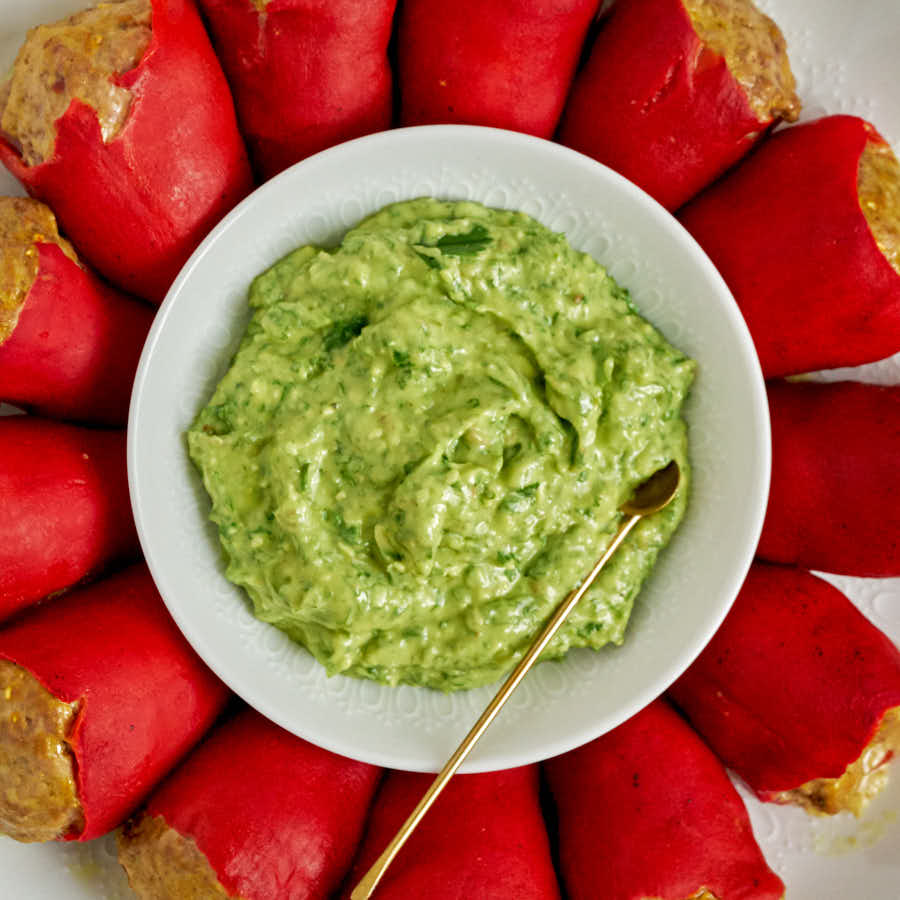 Green avocado sauce in a white bowl surrounded by stuffed red peppers.