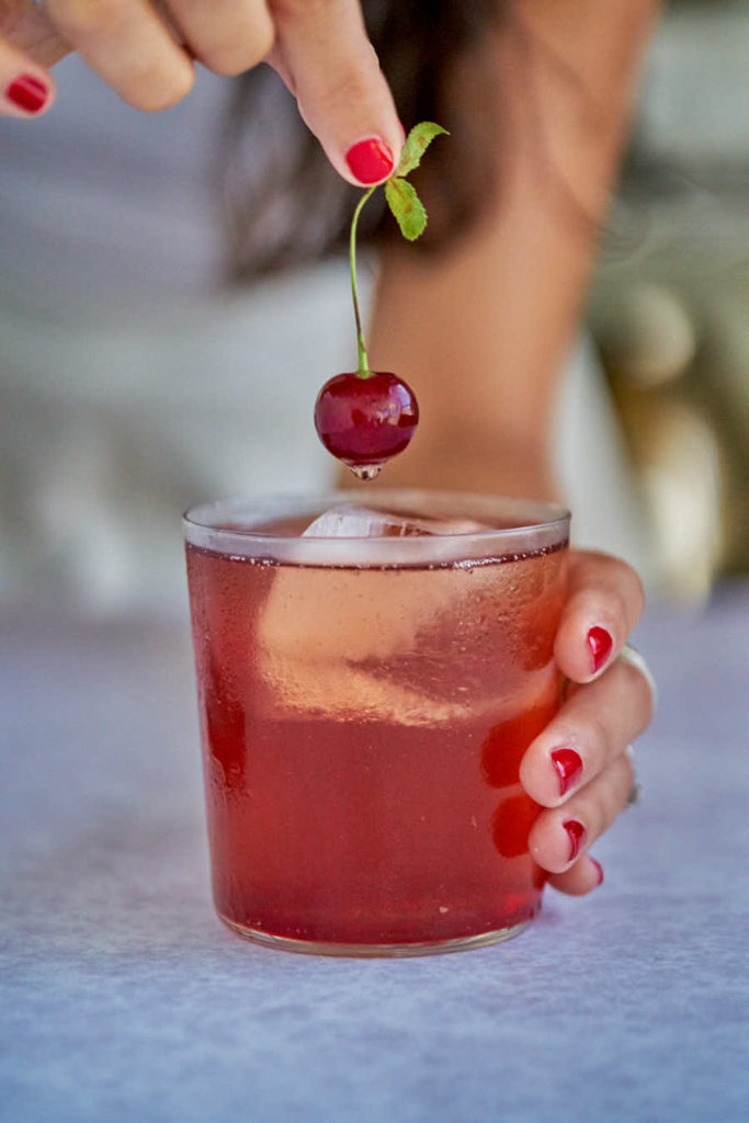 Girl holding dripping cherry over red cocktail.