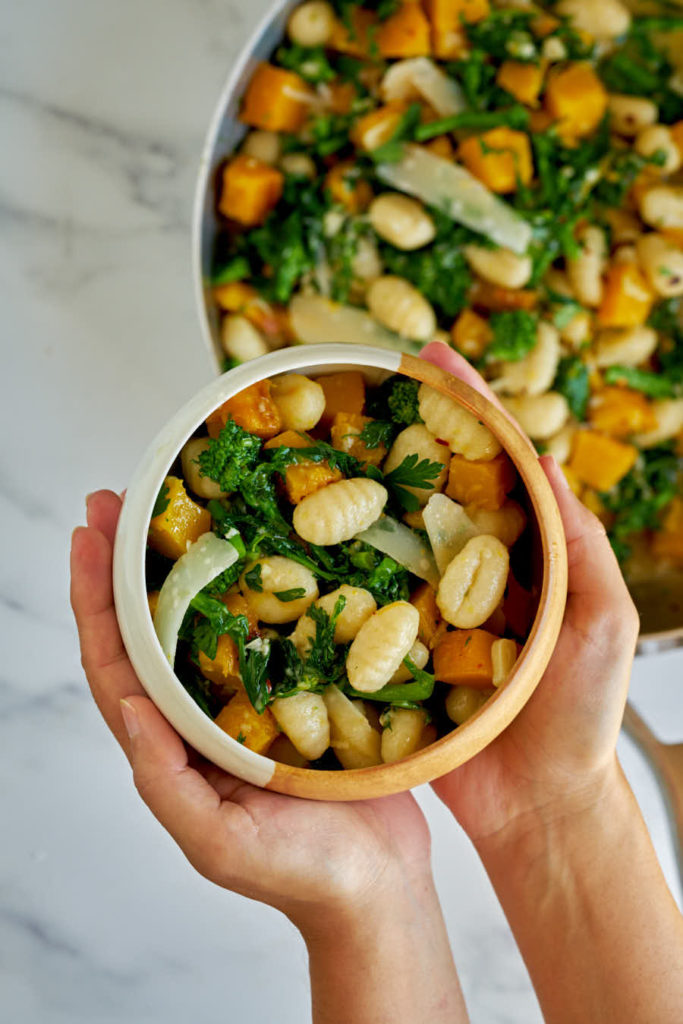 Two hands holding ceramic bowl with vegetables and gnocchi.