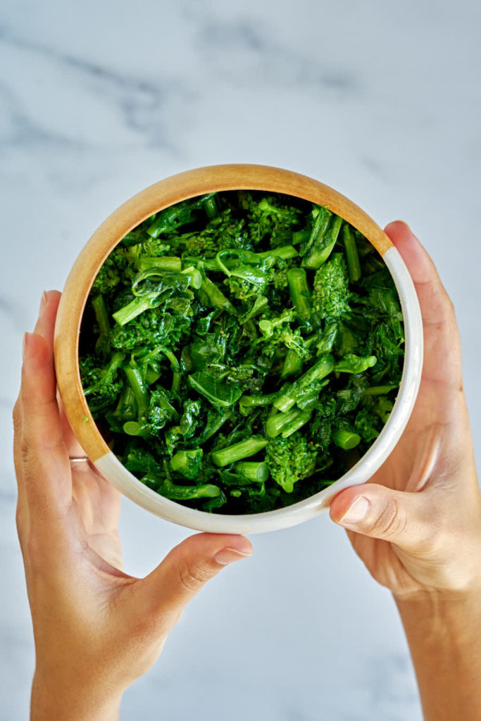 Two hands holding ceramic bowl with cooked green broccoli rabe.