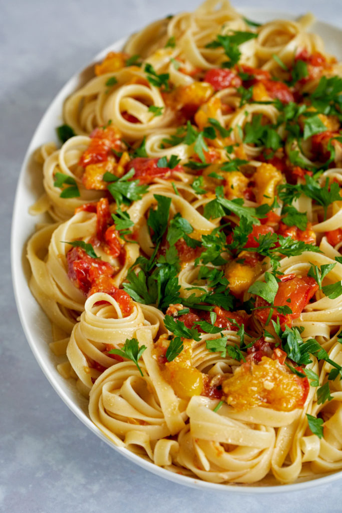 White platter with twirled pasta, topped with confit tomatoes and parsley.