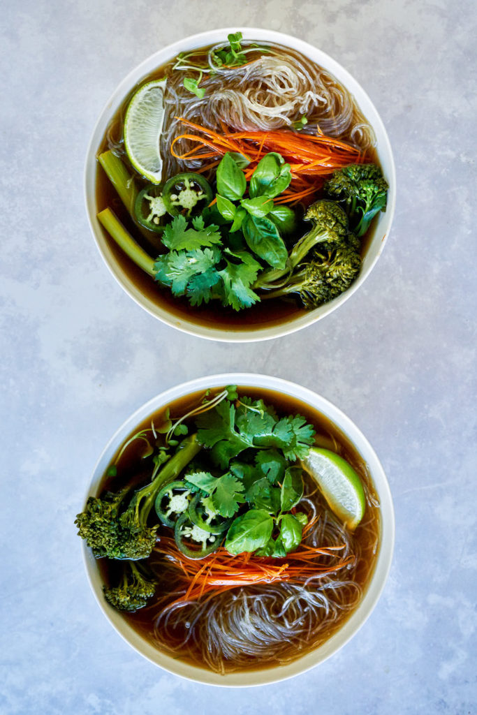 Two bowls of noodle soup with vegetables and herbs.